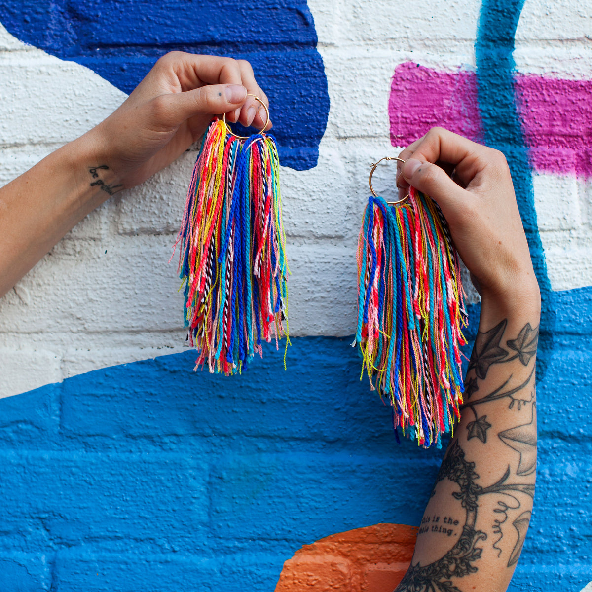 Person holding colorful tassel earrings against a multicolored mural.