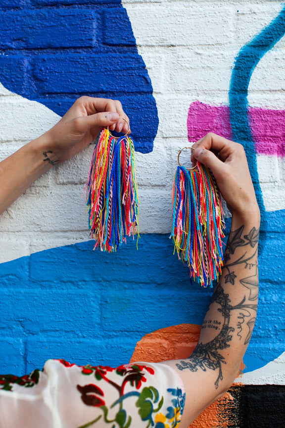 Person holding colorful tassel earrings against a multicolored mural.