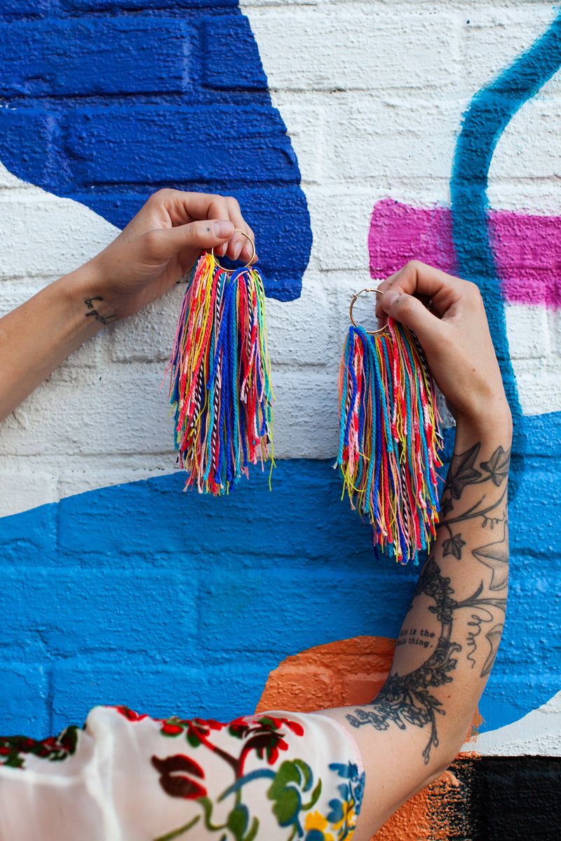 Person holding colorful tassel earrings against a multicolored mural.