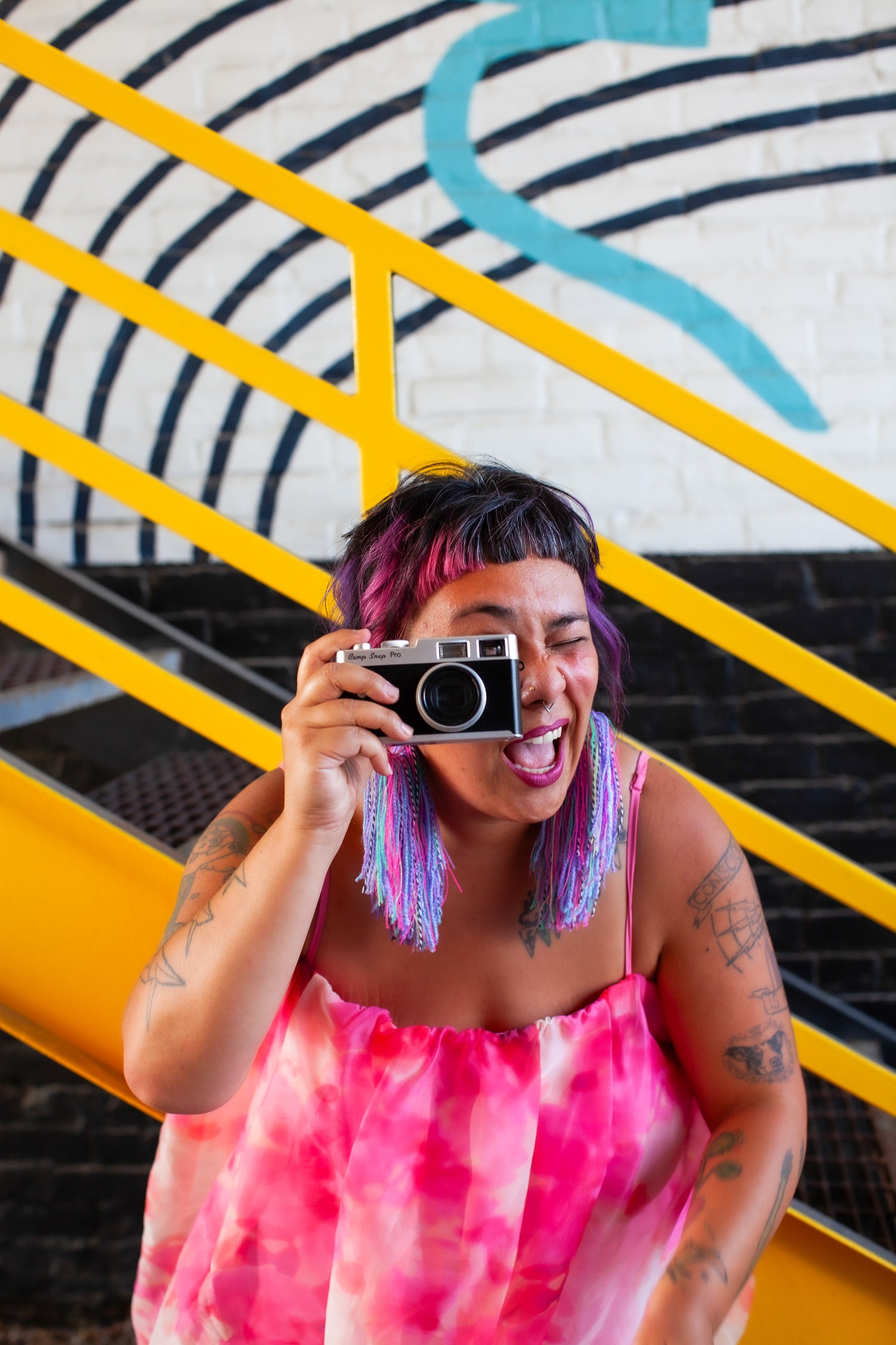 Person holding a camera in front of a colorful wall with yellow steps and bold colorful earrings